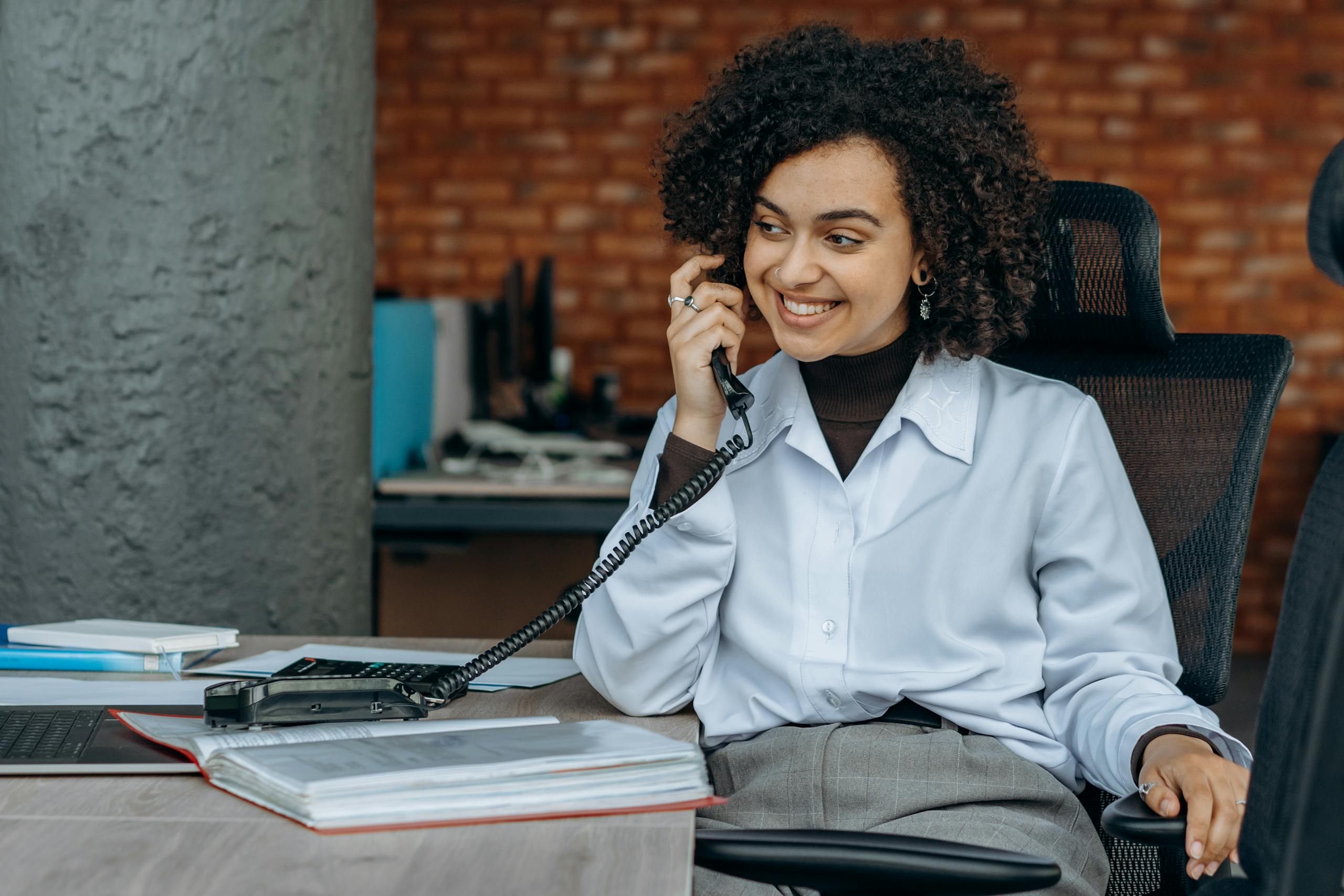 Business woman with curly hair smiles while taking a call in an office environment.
